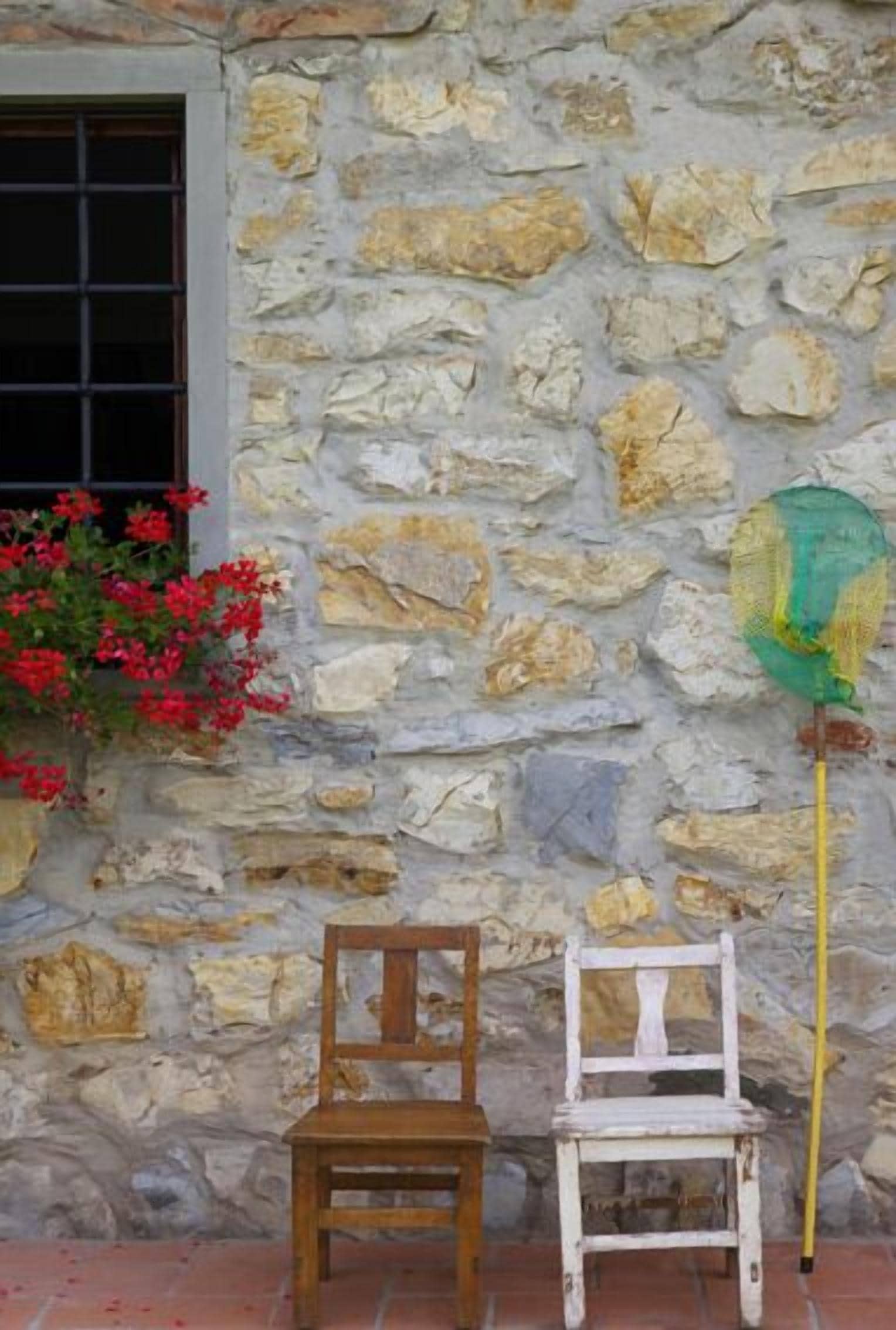 A rustic stone wall with a window, a wooden chair, a white chair, and a green net hanging on the wall.