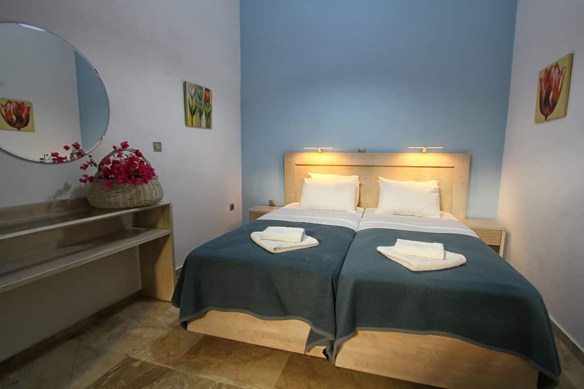 A double bed with two white pillows and a dark blue bedspread, a round mirror, a wicker basket with pink flowers, and a small nightstand with a lamp.