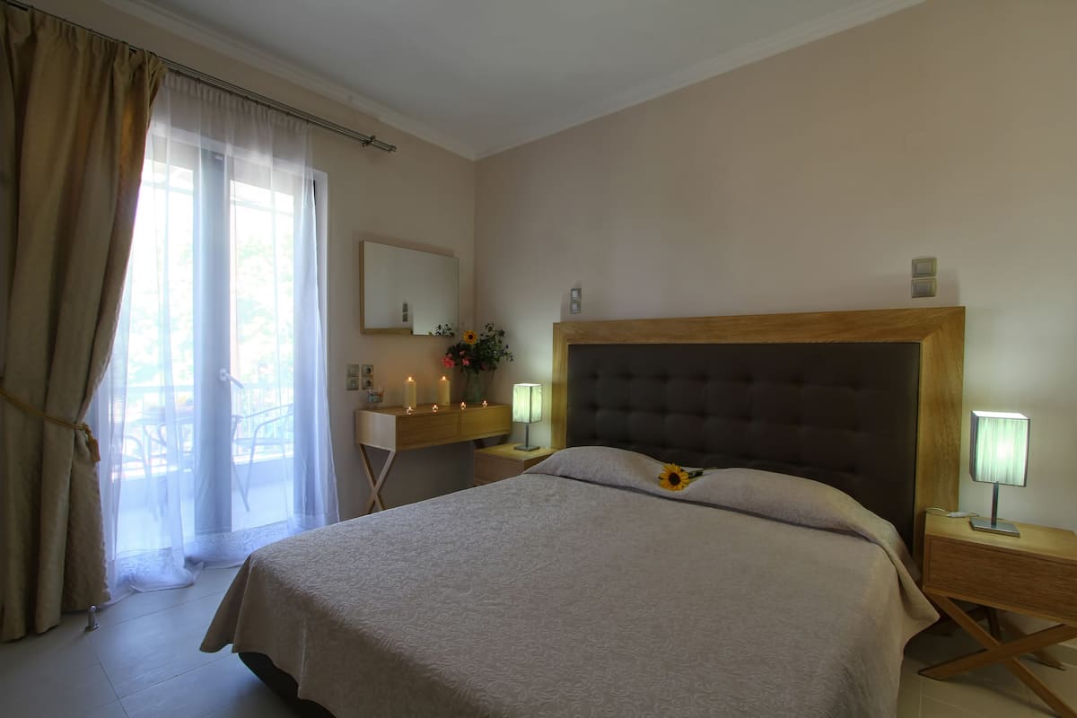A bedroom with a large bed, bedside lamps, a nightstand, a dresser, and a window with sheer curtains.