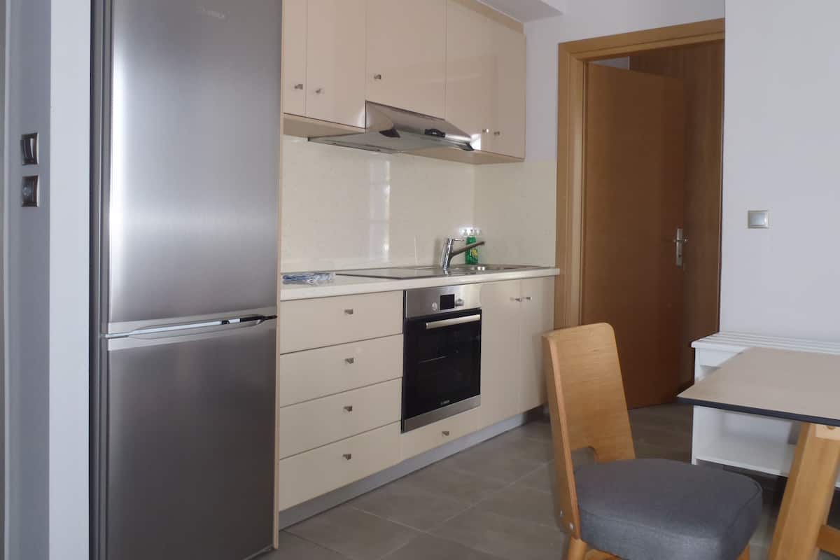 A compact kitchen with a stainless steel refrigerator, a built-in oven, and a microwave. There is a wooden chair and a small table with a chair. The kitchen has beige cabinets and a white countertop.