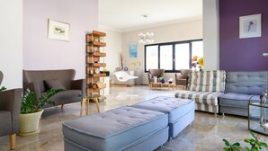 A modern living room with a grey sofa, a striped sofa, and a blue ottoman. There is a wooden shelf unit and a white chair. - Ilioxenia Studio & Apartments (Chios)