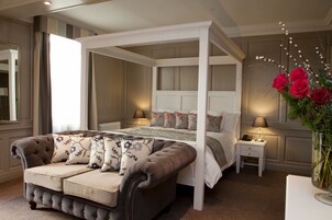 A four-poster canopy bed with a tufted headboard, a tufted sofa with floral cushions, and a bedside table with a lamp.