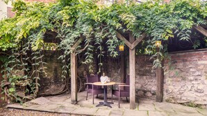 Un espacio al aire libre cubierto con una mesa y sillas, rodeado de plantas trepadoras y un muro de piedra.