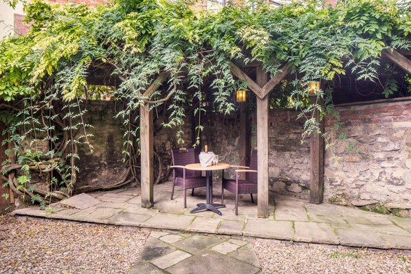 A covered outdoor seating area with a table and chairs, surrounded by climbing plants and a stone wall.