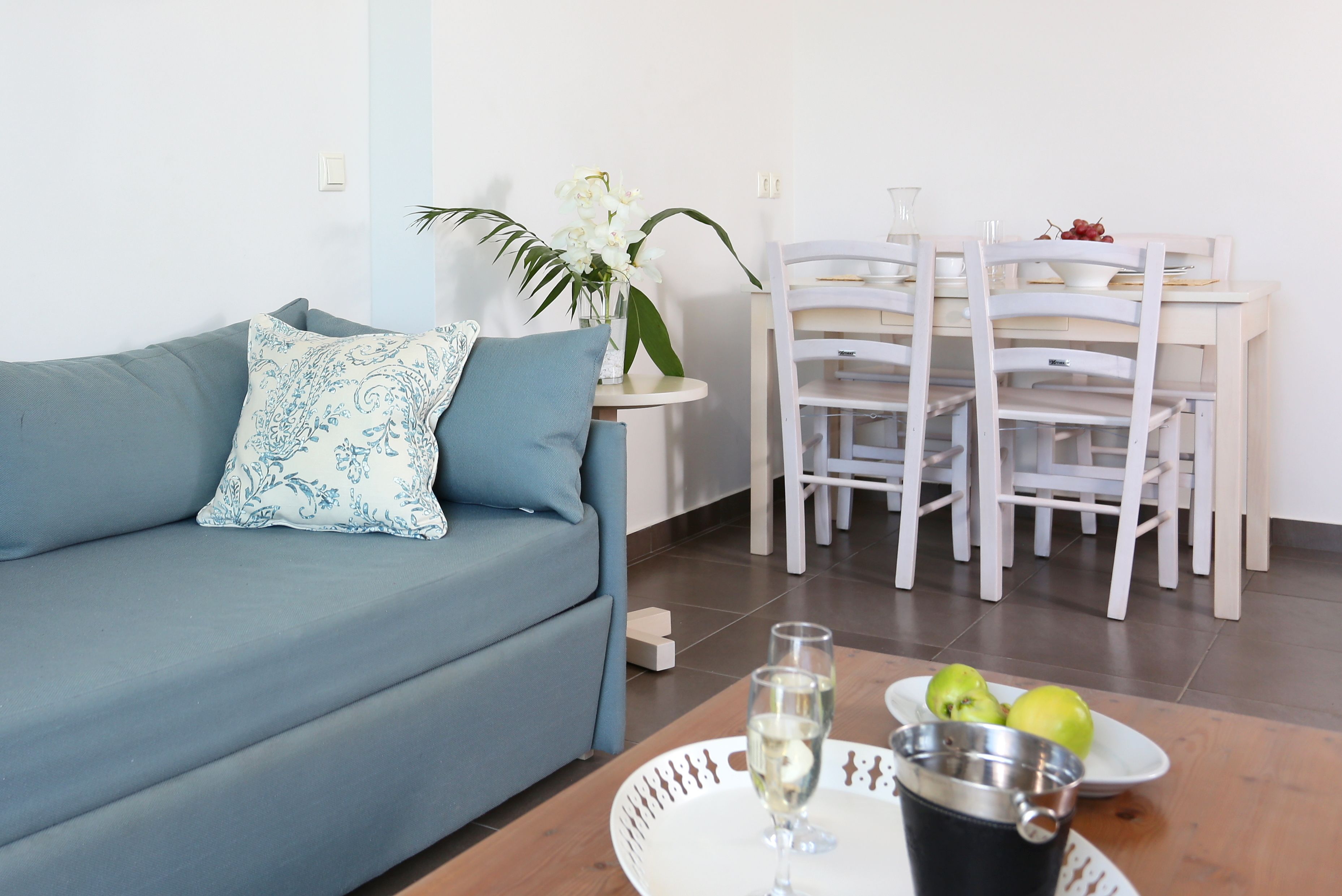 A living room with a blue sofa, a small table with a glass of white wine, a metal pitcher, and a plate with green apples.