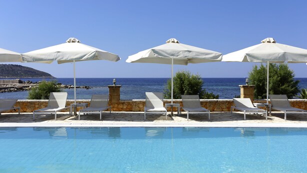 Zona de piscina amb gandules blanques i ombrel·les, cel blau clar i vista al mar.
