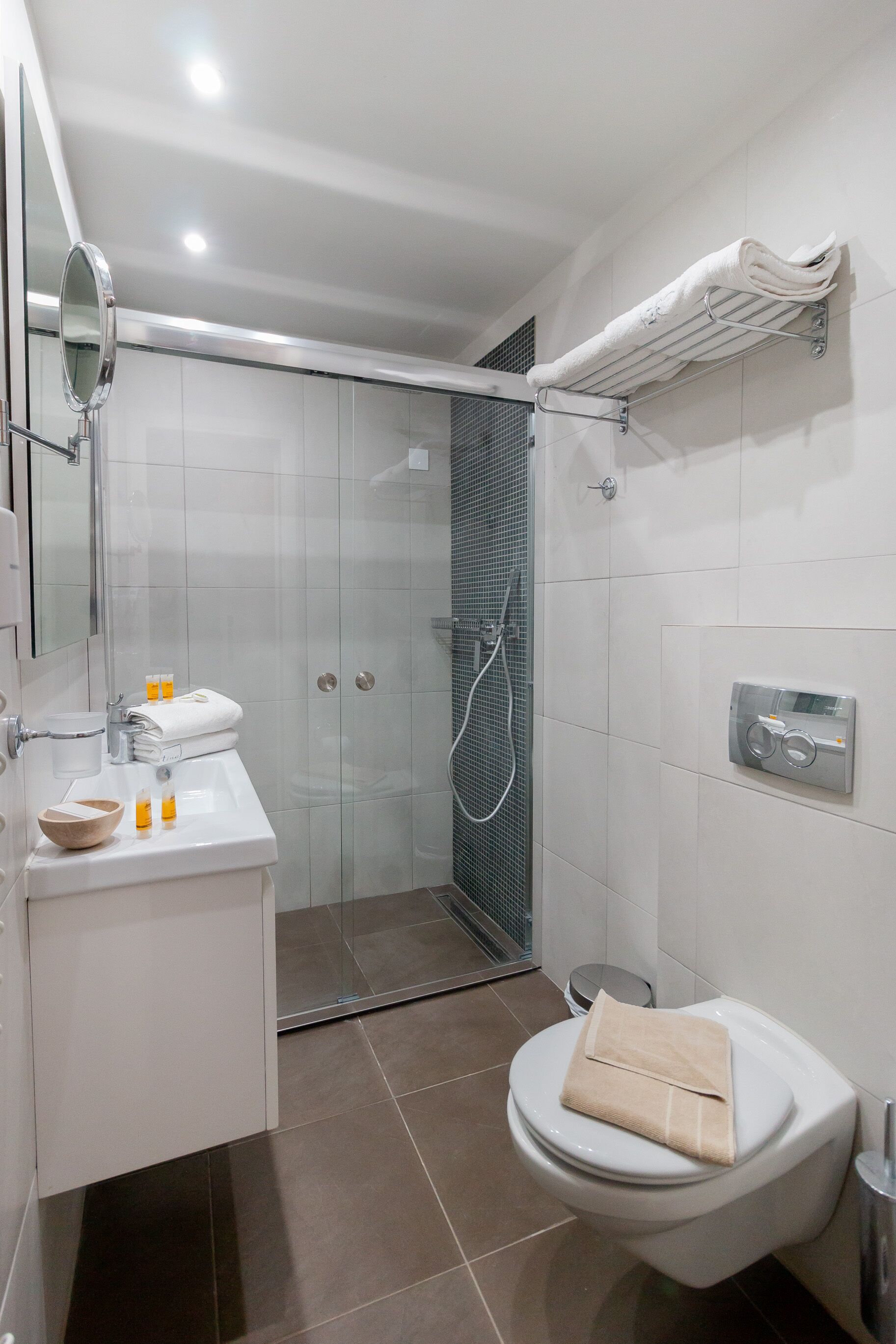 Un baño moderno con cabina de ducha de vidrio, inodoro con toalla color beige, lavamanos con artículos de aseo y espejo.