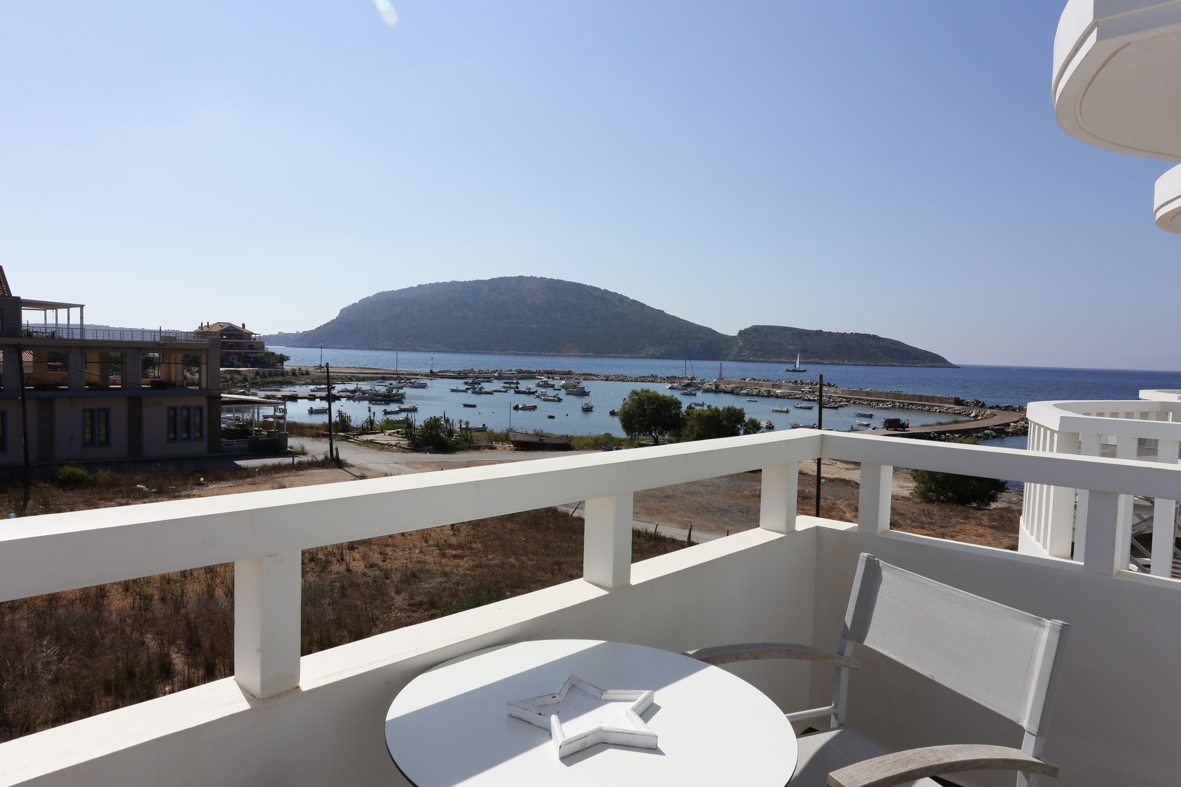 A balcony with a table and chairs, overlooking a marina with boats and a hilly landscape.