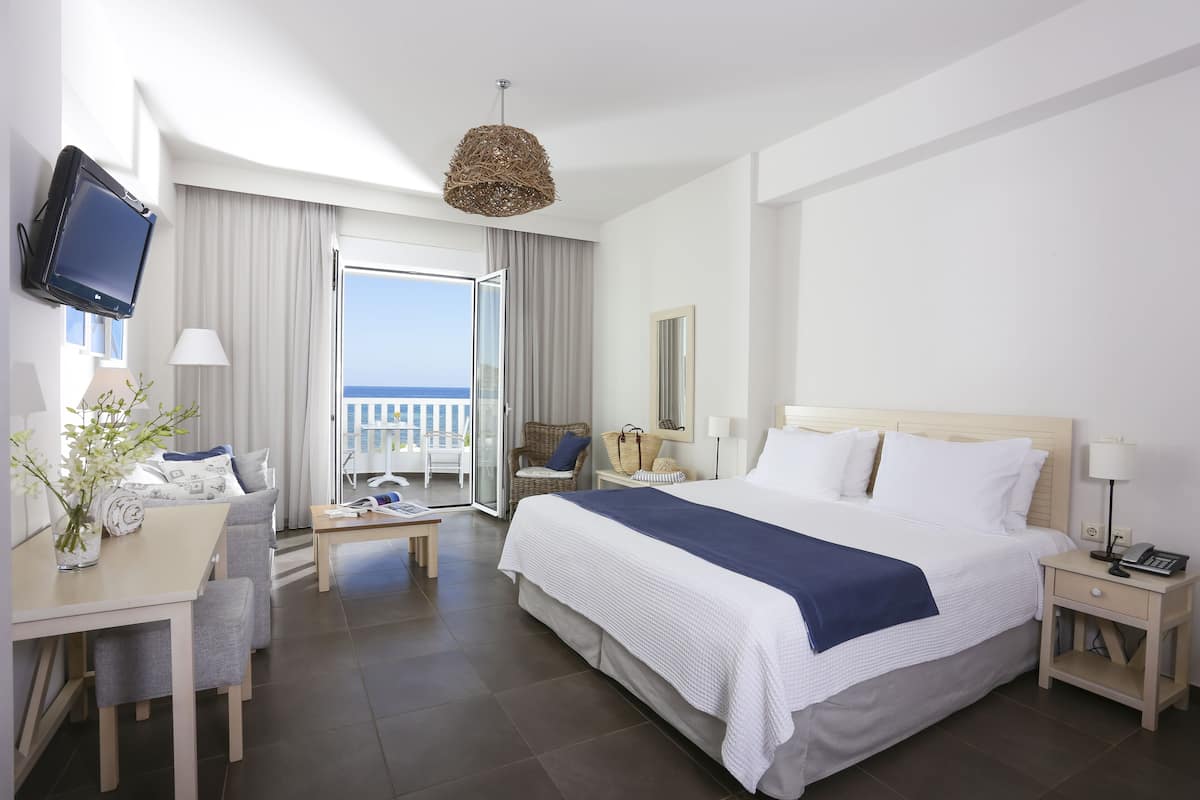 A hotel room with a large bed, a desk with a chair, a television mounted on the wall, and a balcony with a view of the sea.