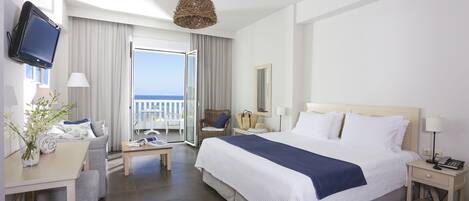 Habitación de hotel con una cama grande, un escritorio con silla, un televisor montado en la pared y un balcón con vistas al mar.