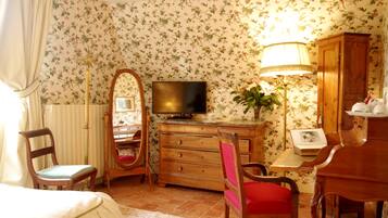 A room with floral wallpaper, a wooden dresser, a television, a desk with a lamp, and a chair with a red cushion.