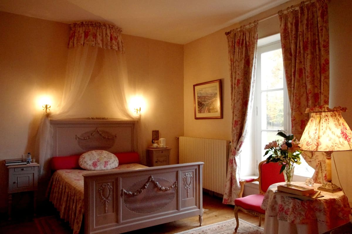 Ein Schlafzimmer mit Bett, Nachttisch, Lampe, Stuhl und einem Fenster mit Vorhängen.