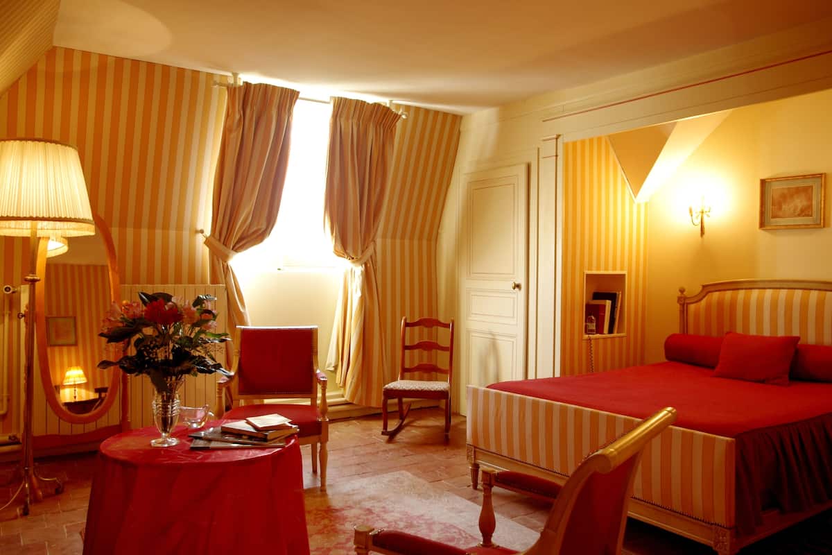 Ein Hotelzimmer mit einem Bett, einem Sessel, einem Tisch mit Blumen und einer Lampe, sowie Vorhängen.