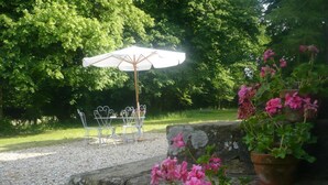 Een tuinpatio met een tafel en stoelen onder een witte parasol, omgeven door groen en potplanten.