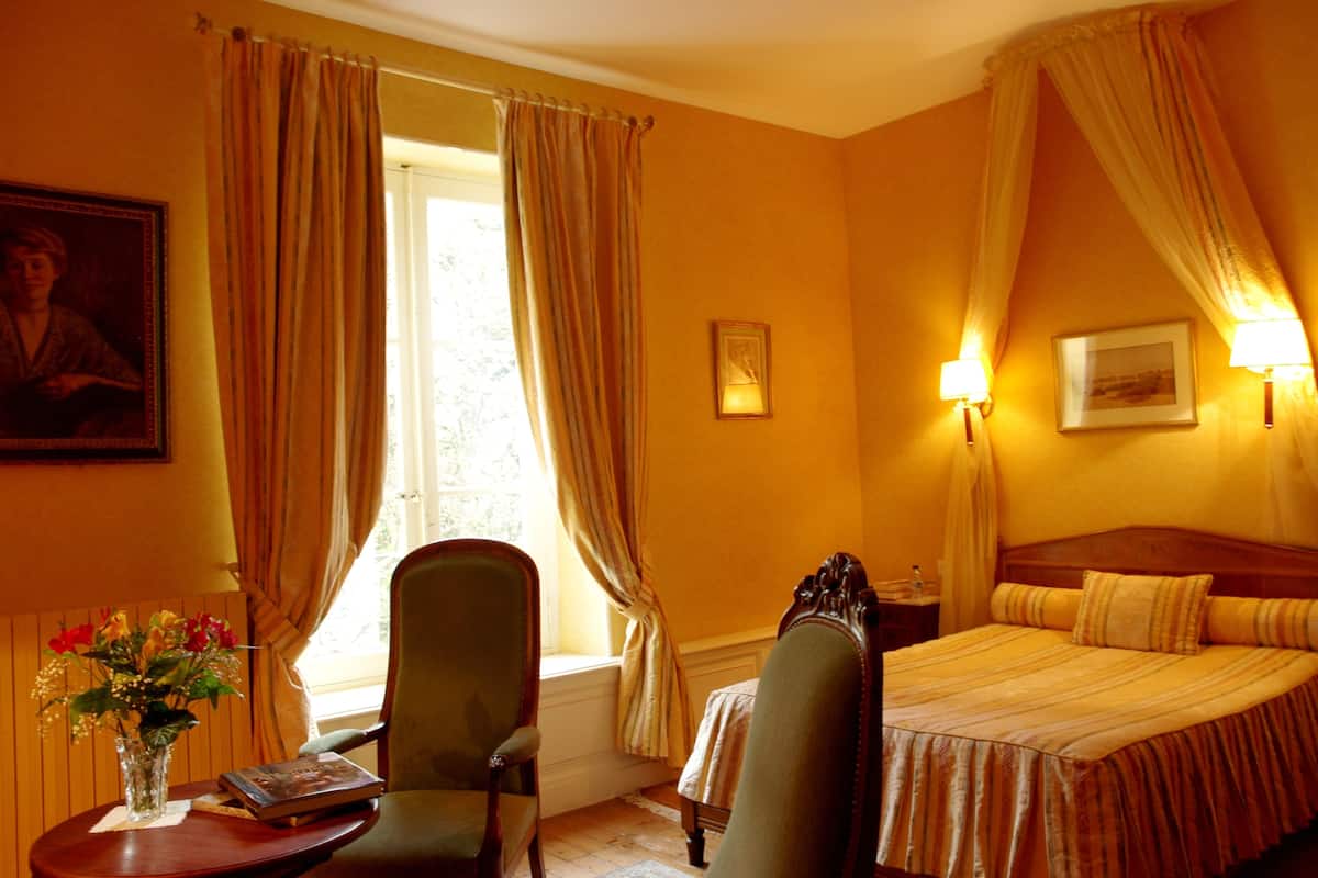 Ein Hotelzimmer mit Bett, Sessel, Tisch mit Blumenvase und einem fenster mit Vorhängen.