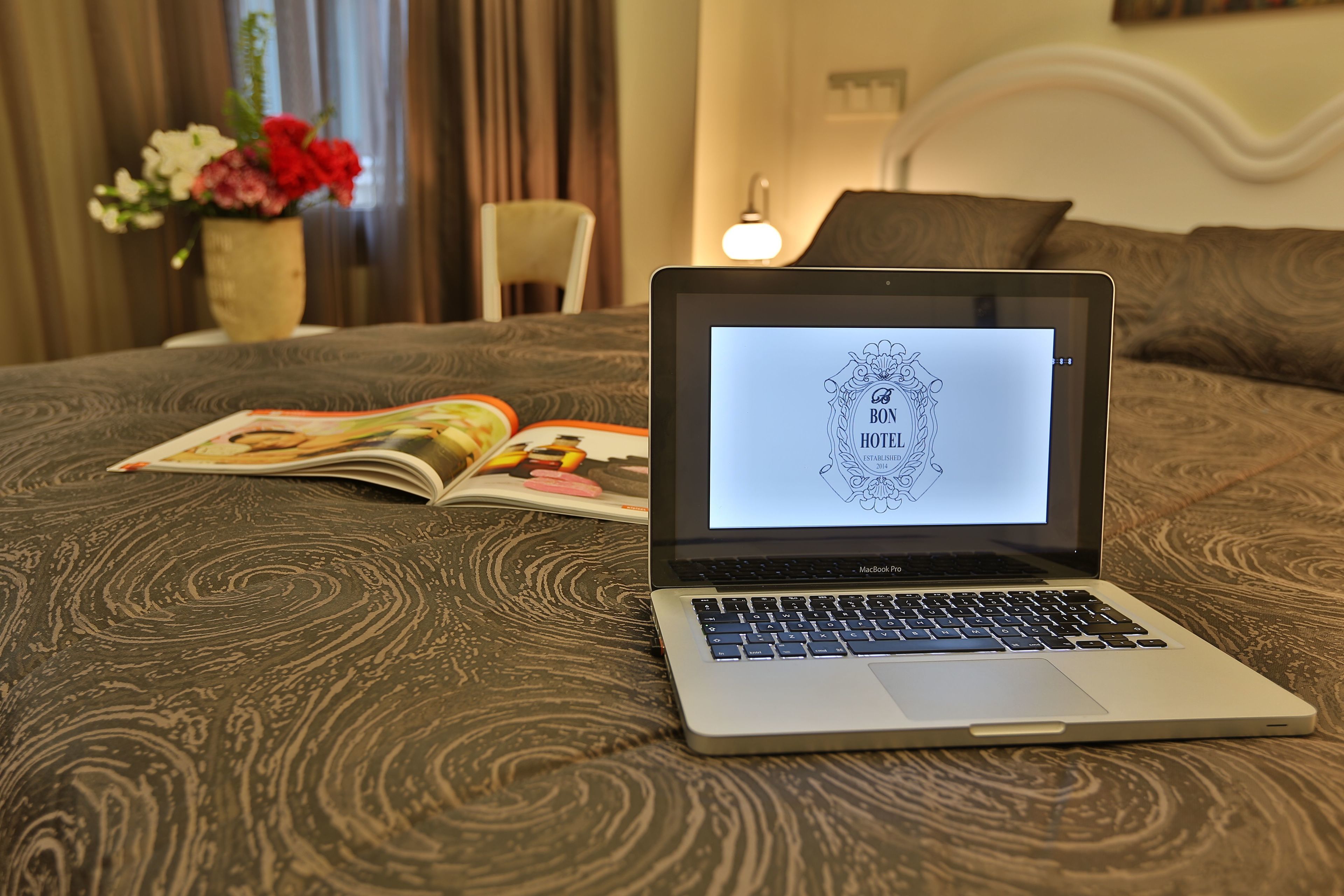 A MacBook on a bed with a magazine, a vase of flowers, and a lamp in the background.