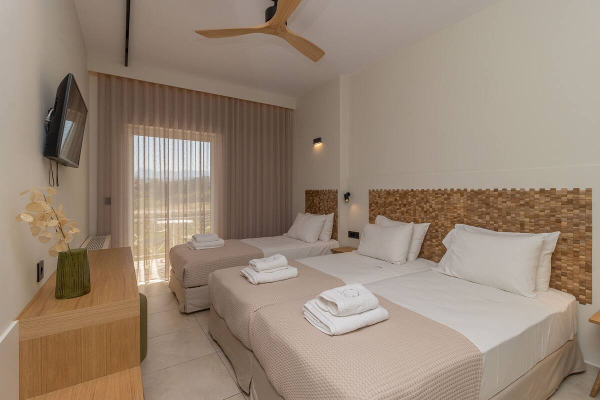 A hotel room with two beds, a TV mounted on the wall, a ceiling fan, and a large window with curtains.