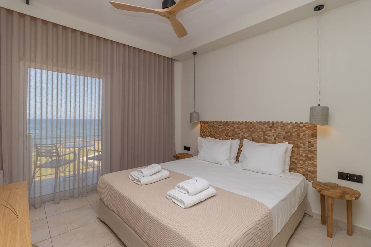 A hotel room with a large bed, a ceiling fan, a view of the sea, and a table with chairs outside the window.