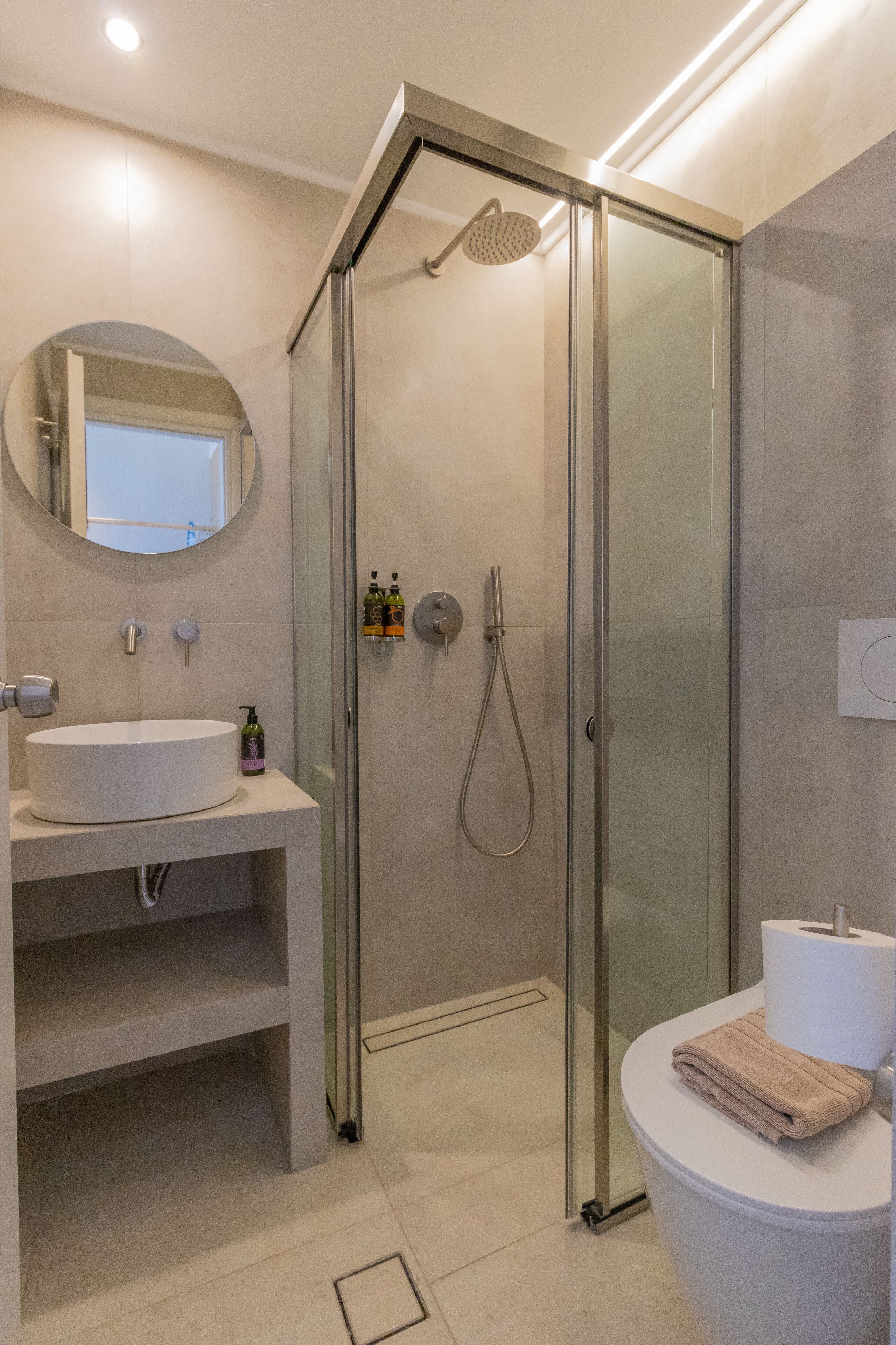 A modern bathroom with a glass-enclosed shower, a round mirror, a white sink, and a toilet with a towel.