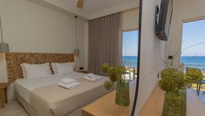 Quarto de hotel com uma cama grande, cabeceira de madeira, mesa de cabeceira com abajur, TV fixada na parede e vista para a praia.