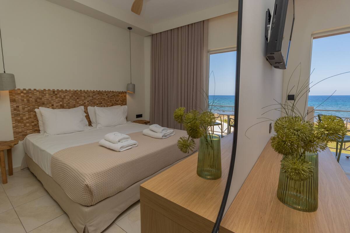 A hotel room with a large bed, a wooden headboard, a bedside table with a lamp, a TV mounted on the wall, and a view of the beach.