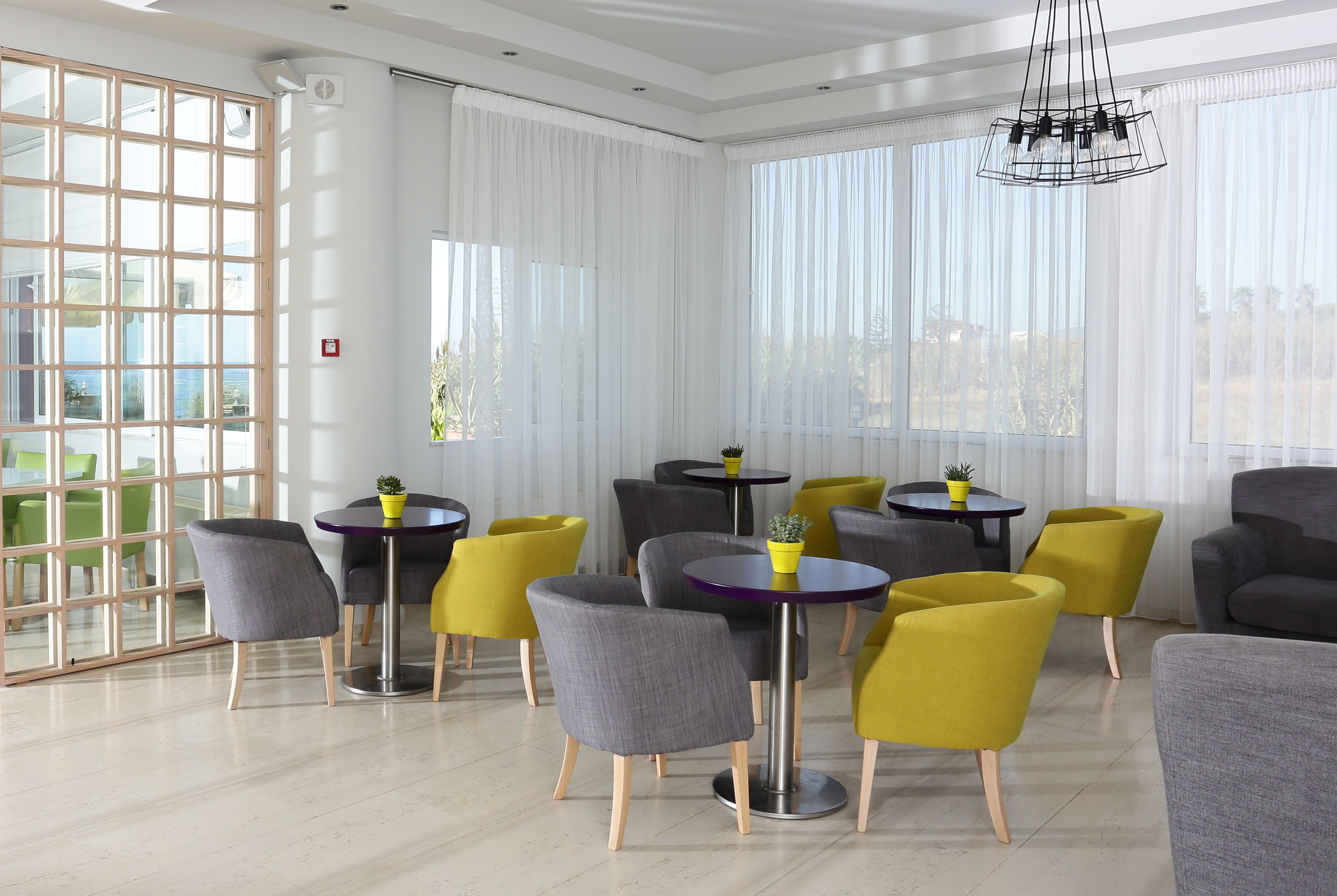 A modern dining area with round tables and chairs, large windows with sheer curtains, and a view of the outdoors.