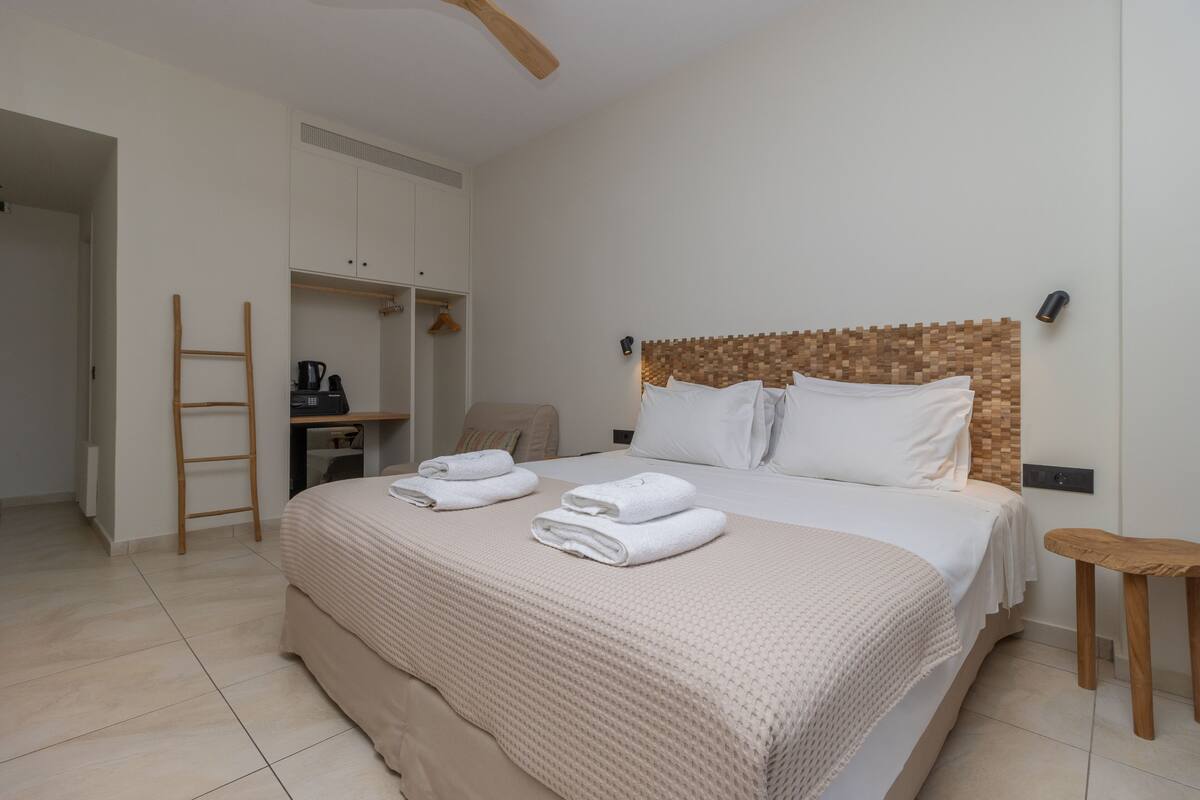 A neatly made bed with a textured beige bedspread and pillows, a wooden ladder, a small wooden stool, and a desk with a kettle.