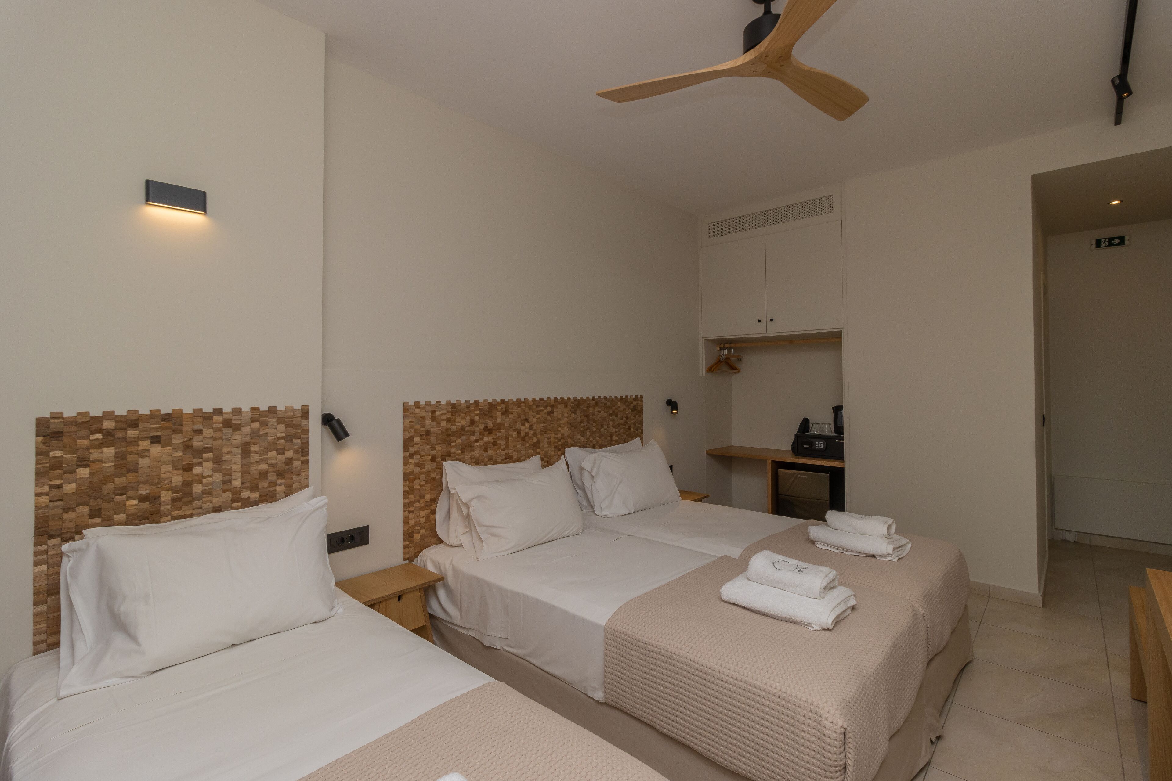 Quarto de hotel com duas camas, uma cabeceira de madeira, um ventilador de teto, uma secretária pequena e um guarda-roupa.