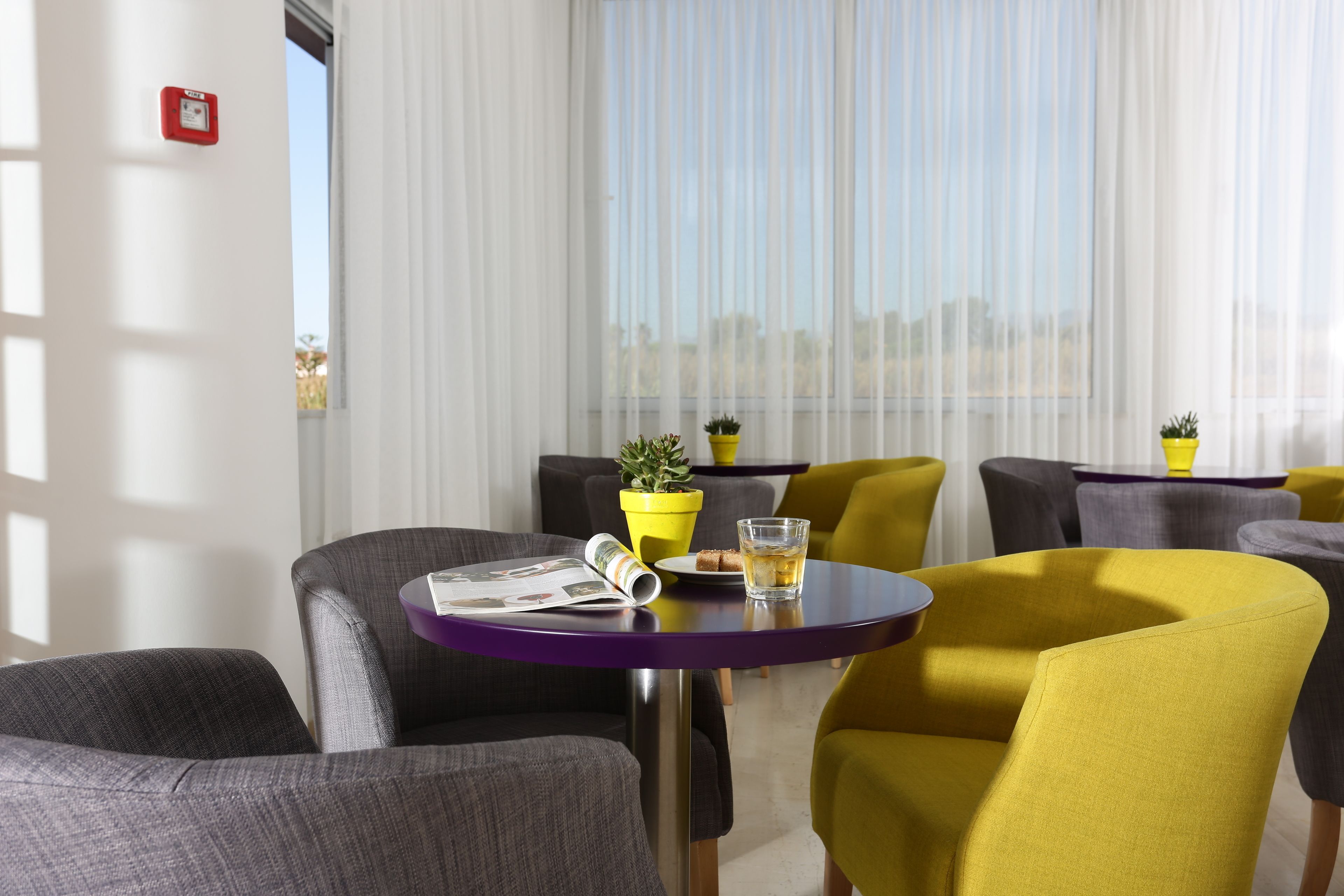 Ein modernes Hotelzimmer mit einem violetten Tisch, gelb-grauen Stühlen und einem Fenster mit transparenten Vorhängen.