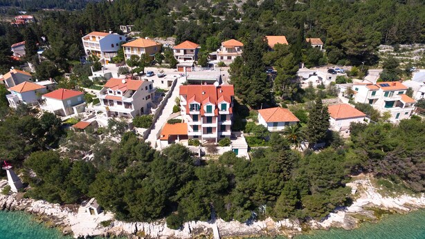 An aerial view of a coastal residential area with houses featuring red-tiled roofs, surrounded by dense greenery and a clear turquoise sea. - Apartments Vala (Vela Luka)