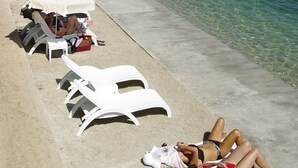 A beach with sun loungers and umbrellas, clear water, and a view of the town in the background. - Apartments Vala (Vela Luka)