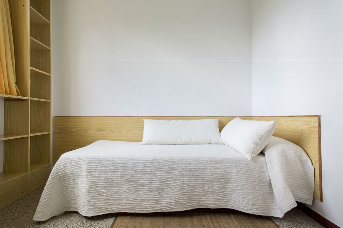 Un lit bien fait, avec une tête de lit en bois, une armoire en bois sur le côté gauche et un mur sobre.