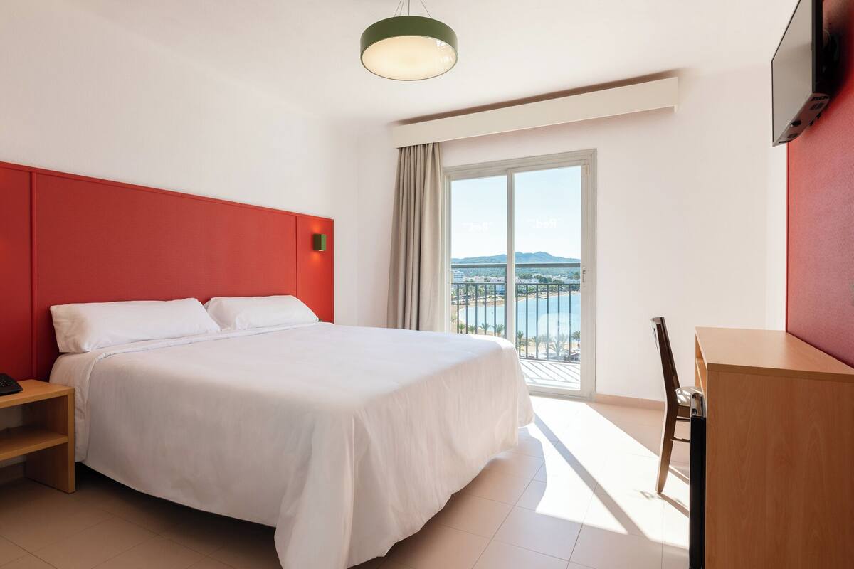 Ein Hotelzimmer mit einem großen Bett, einem Schreibtisch und Blick auf ein Gewässer durch eine Schiebetür.