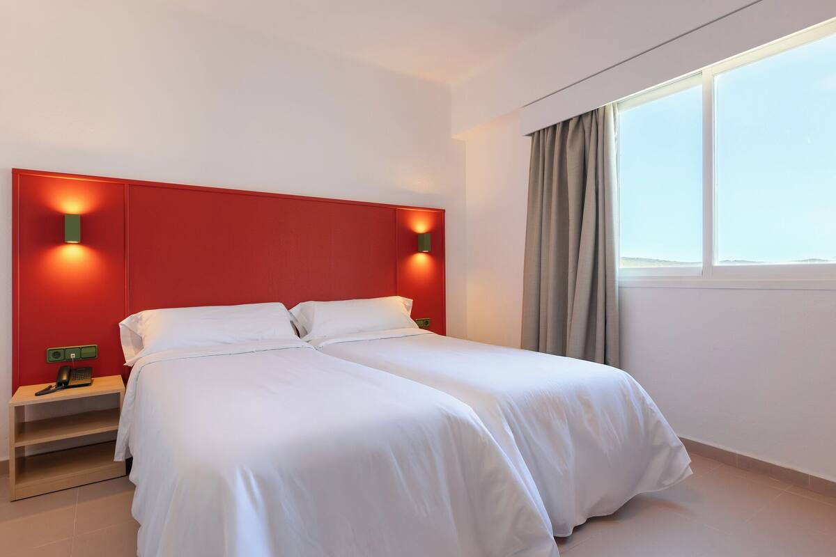 Ein Doppelbett mit weißer Bettwäsche in einem Raum, der durch eine rote Wand hervorgehoben wird, mit zwei Wandleuchten.
