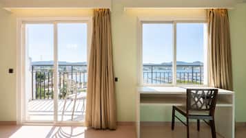 A room with a desk and chair, large windows with curtains, and a view of a body of water and distant mountains.