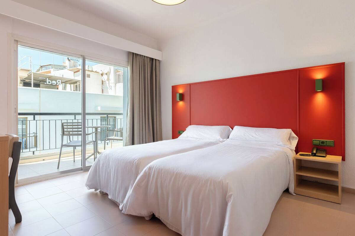 Ein Hotelzimmer mit zwei Betten, einem Balkon mit Blick auf Gebäude und einer roten Akzentwand.