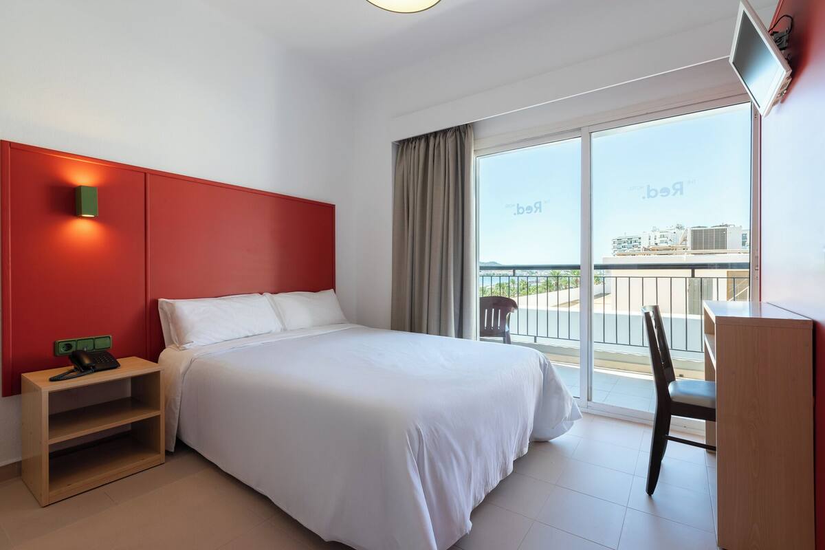 Ein Hotelzimmer mit einem Bett, einem Schreibtisch, einem Stuhl, einem Nachttisch mit Telefon und einem großen Fenster mit Blick auf Gebäude.