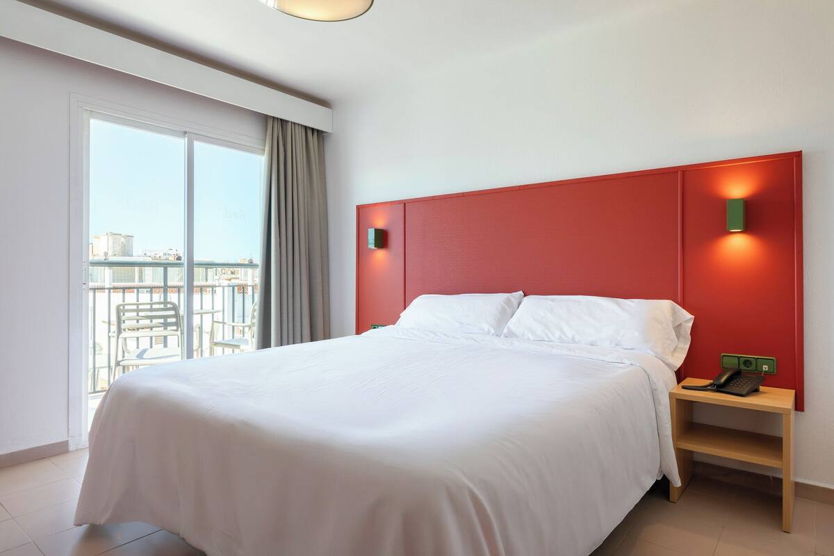 Ein Hotelzimmer mit einem großen Bett, einem Nachttisch mit Telefon und Blick auf die Stadt durch ein großes Fenster.
