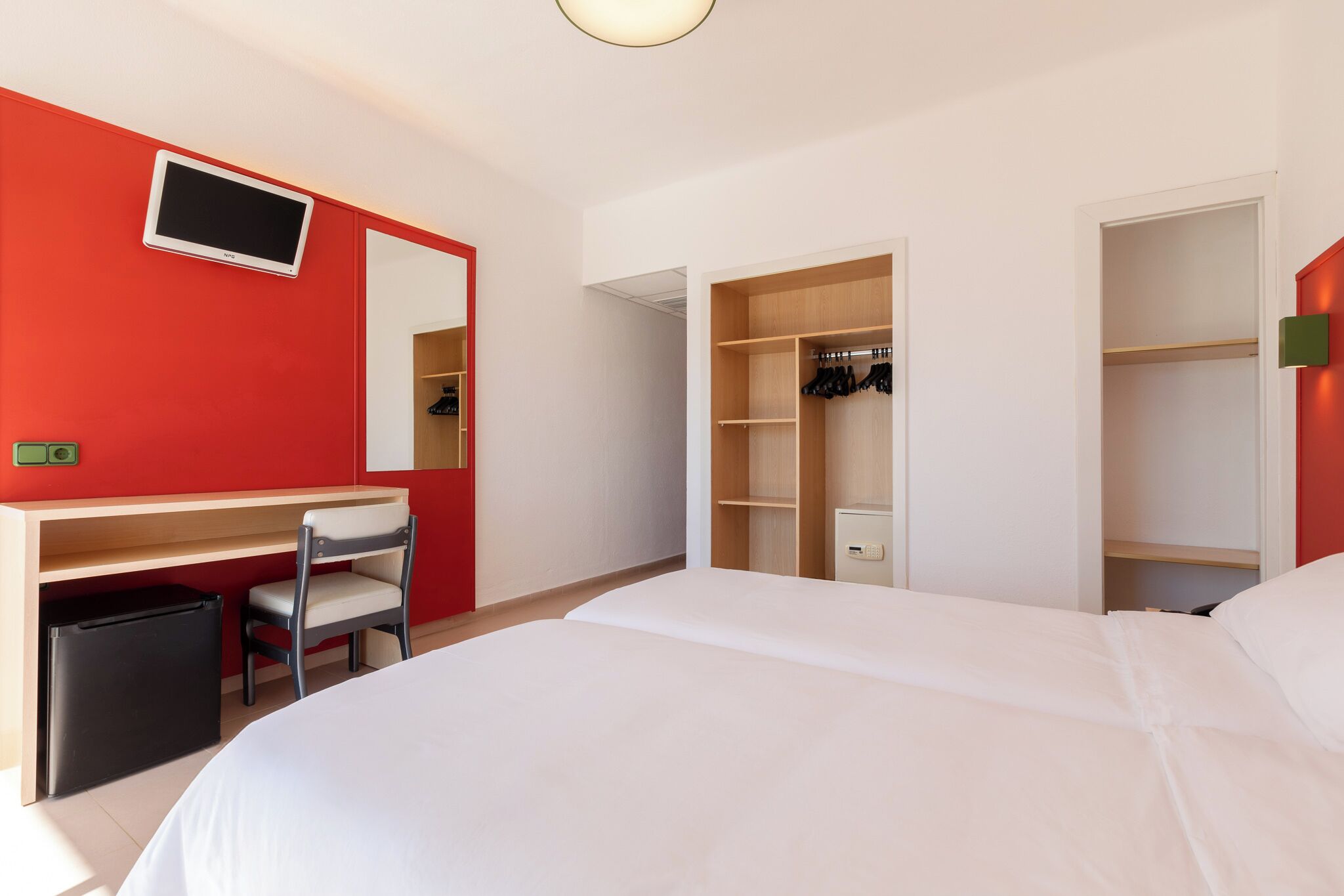 A hotel room with a bed, a desk with a chair, a wardrobe, and a television on the wall.