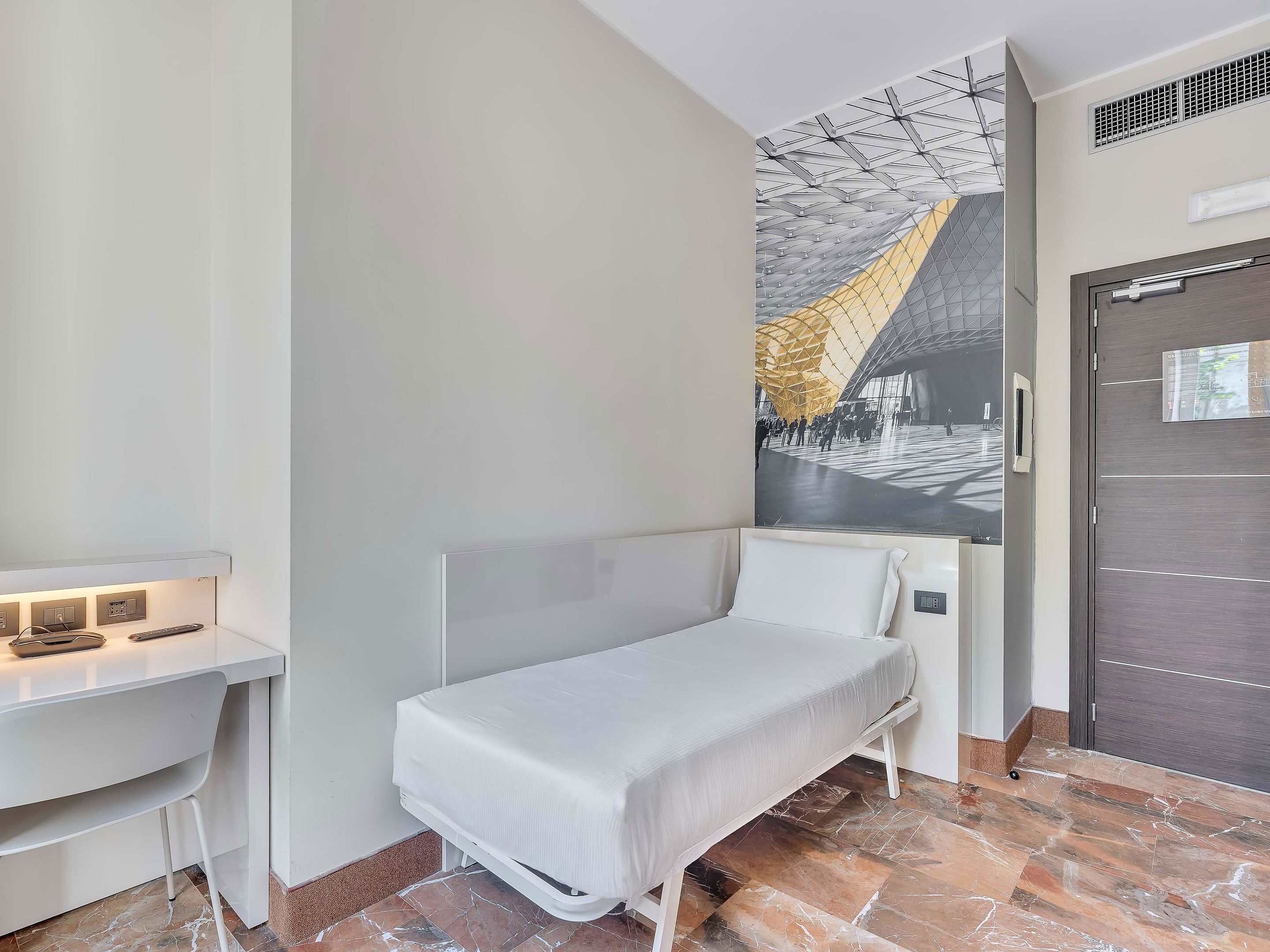 A modern hotel room with a single bed, a desk, and a large window.