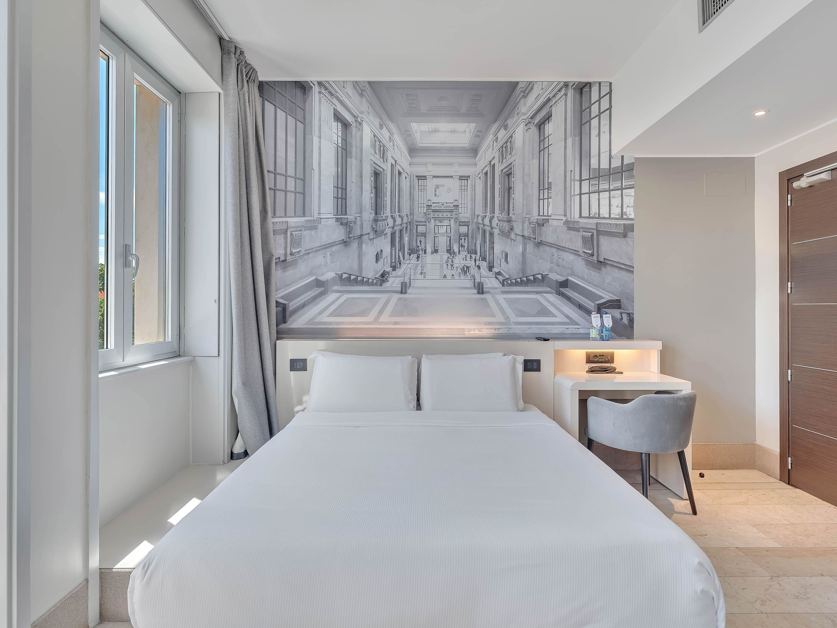 A hotel room with a large bed, a small desk with a chair, a window with curtains, and a mural of a building on the wall.