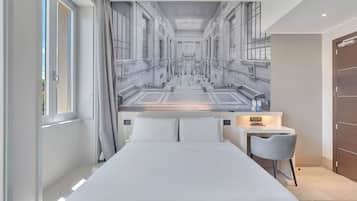 A hotel room with a large bed, a small desk with a chair, a window with curtains, and a mural of a building on the wall.