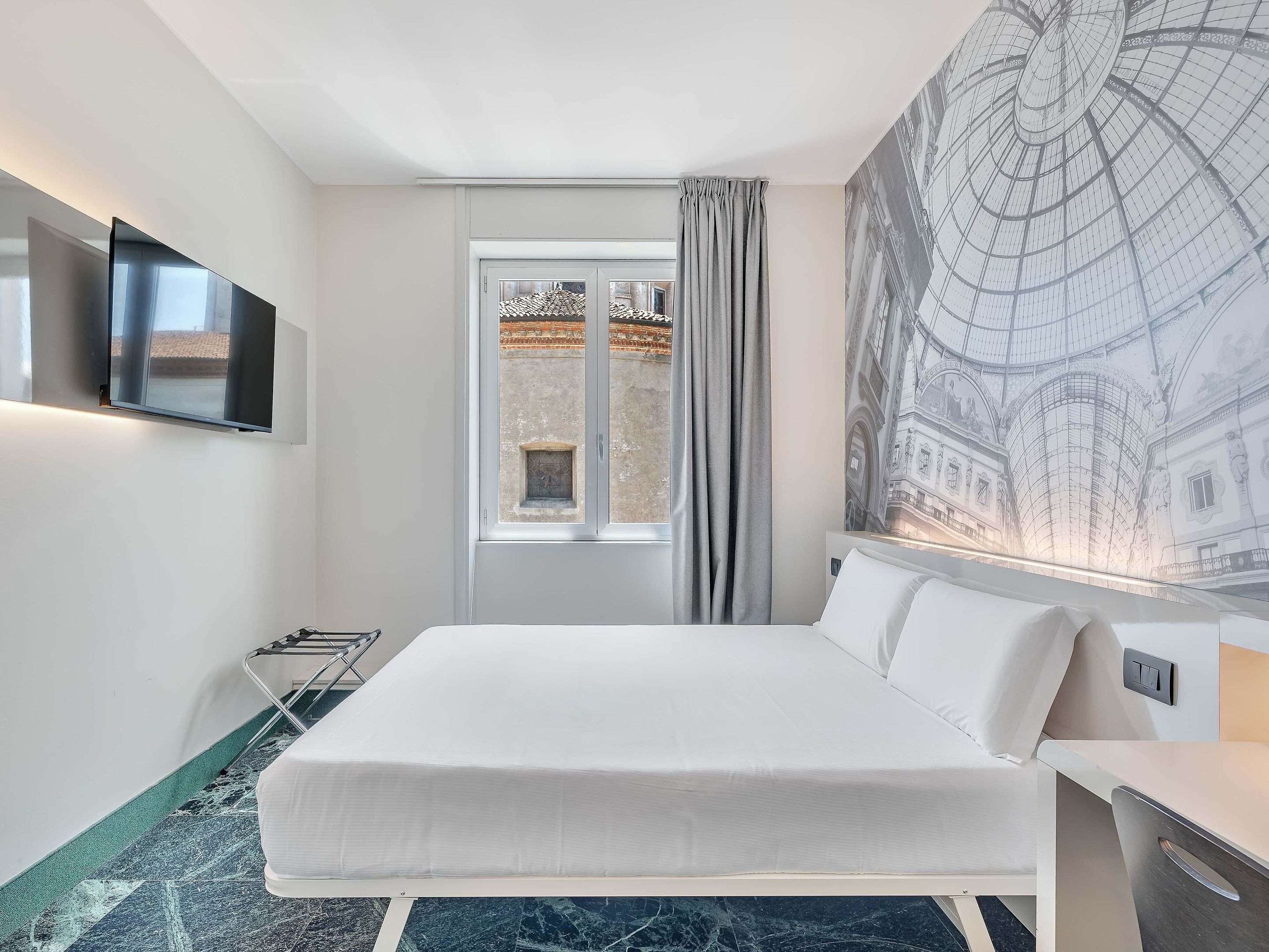 A hotel room with a bed, a TV mounted on the wall, a window with curtains, and a large architectural mural on the wall.