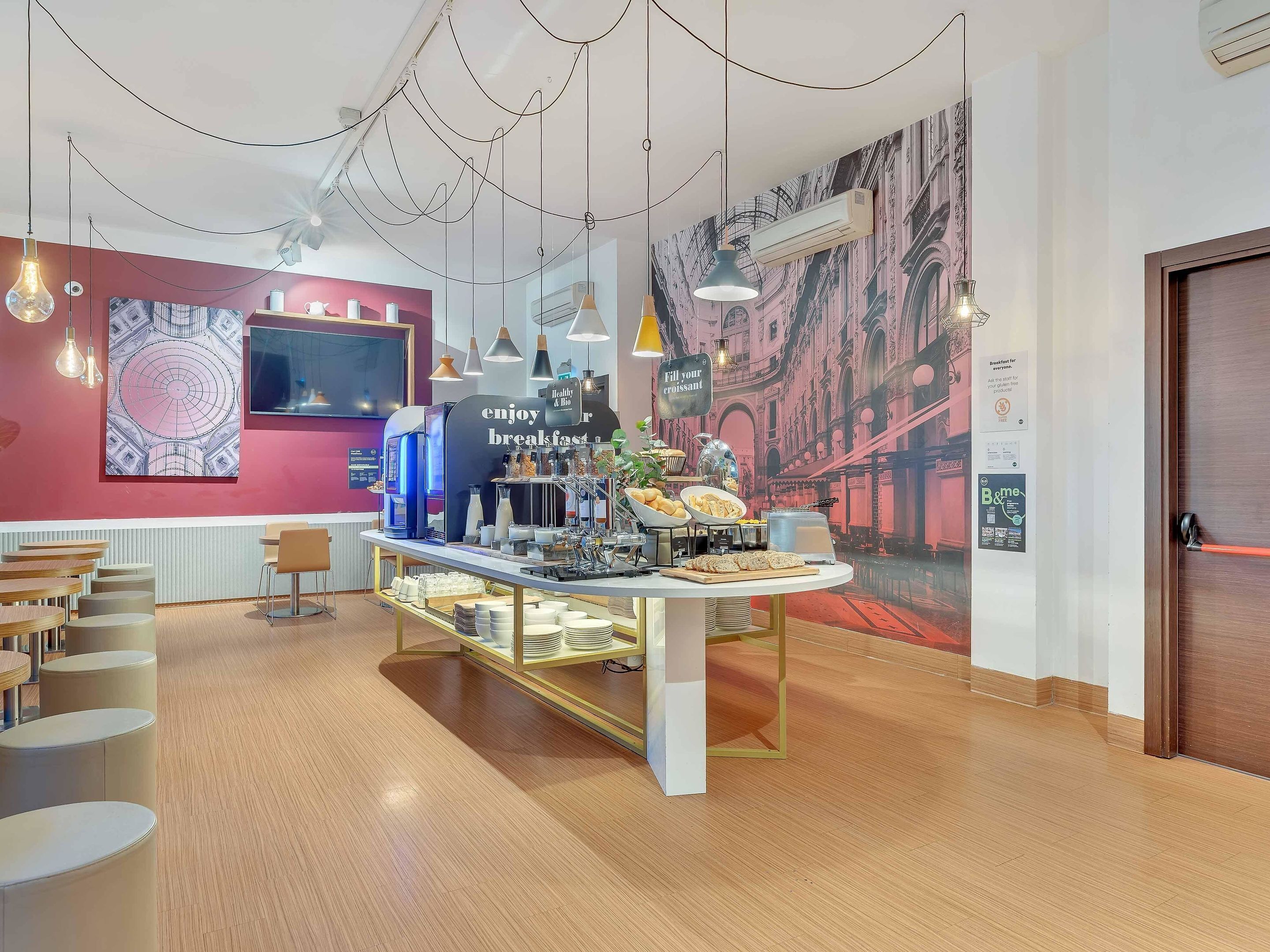 Un espace réservé au petit-déjeuner, avec un comptoir où sont exposées des pâtisseries et des boissons, un coin salon avec des tables, et une fresque représentant un paysage urbain sur le mur.