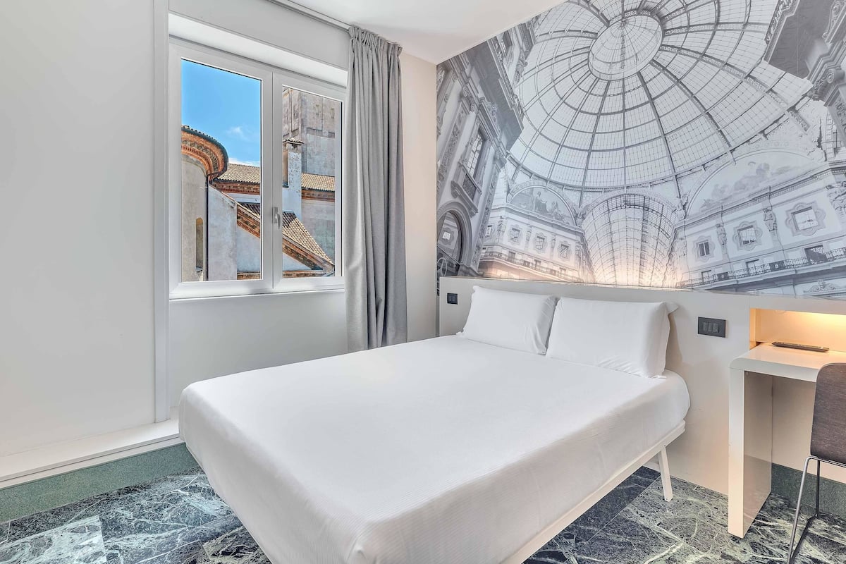 A hotel room with a large bed, a window with a view of a building, and a mural of a historical building on the wall.