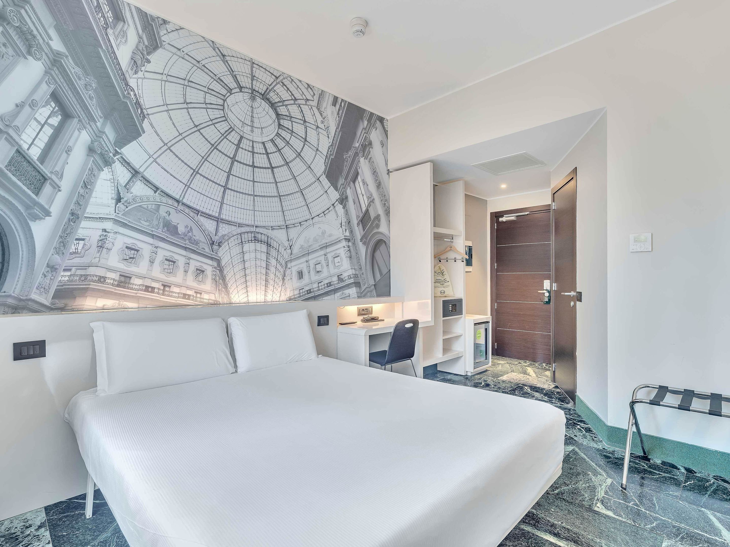 A hotel room with a large bed, a desk, and a chair. There is a mural on the wall depicting a historical building.