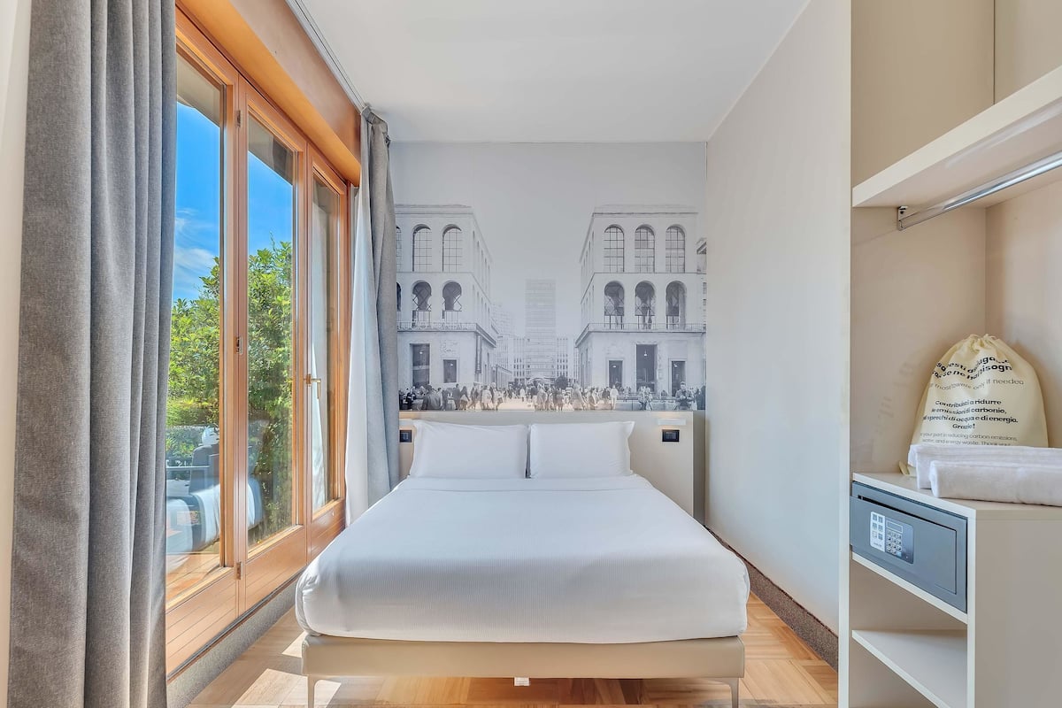 A hotel room with a large bed, a view of a cityscape through the window, and a small bedside table with a lamp.
