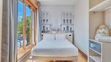 A hotel room with a large bed, a view of a cityscape through the window, and a small bedside table with a lamp.