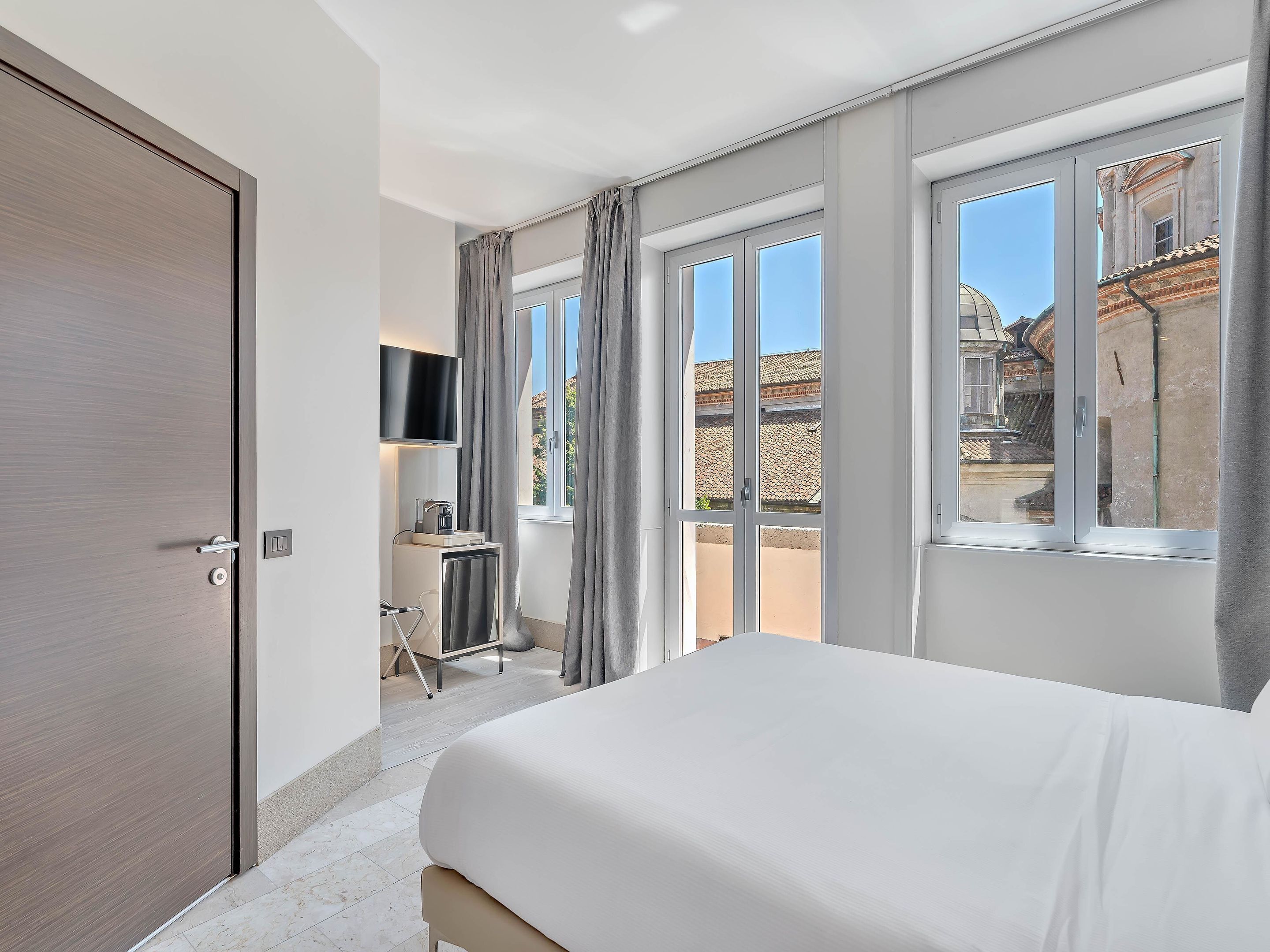 Habitación de hotel con cama, escritorio con silla, televisor montado en la pared y ventanas amplias con cortinas.