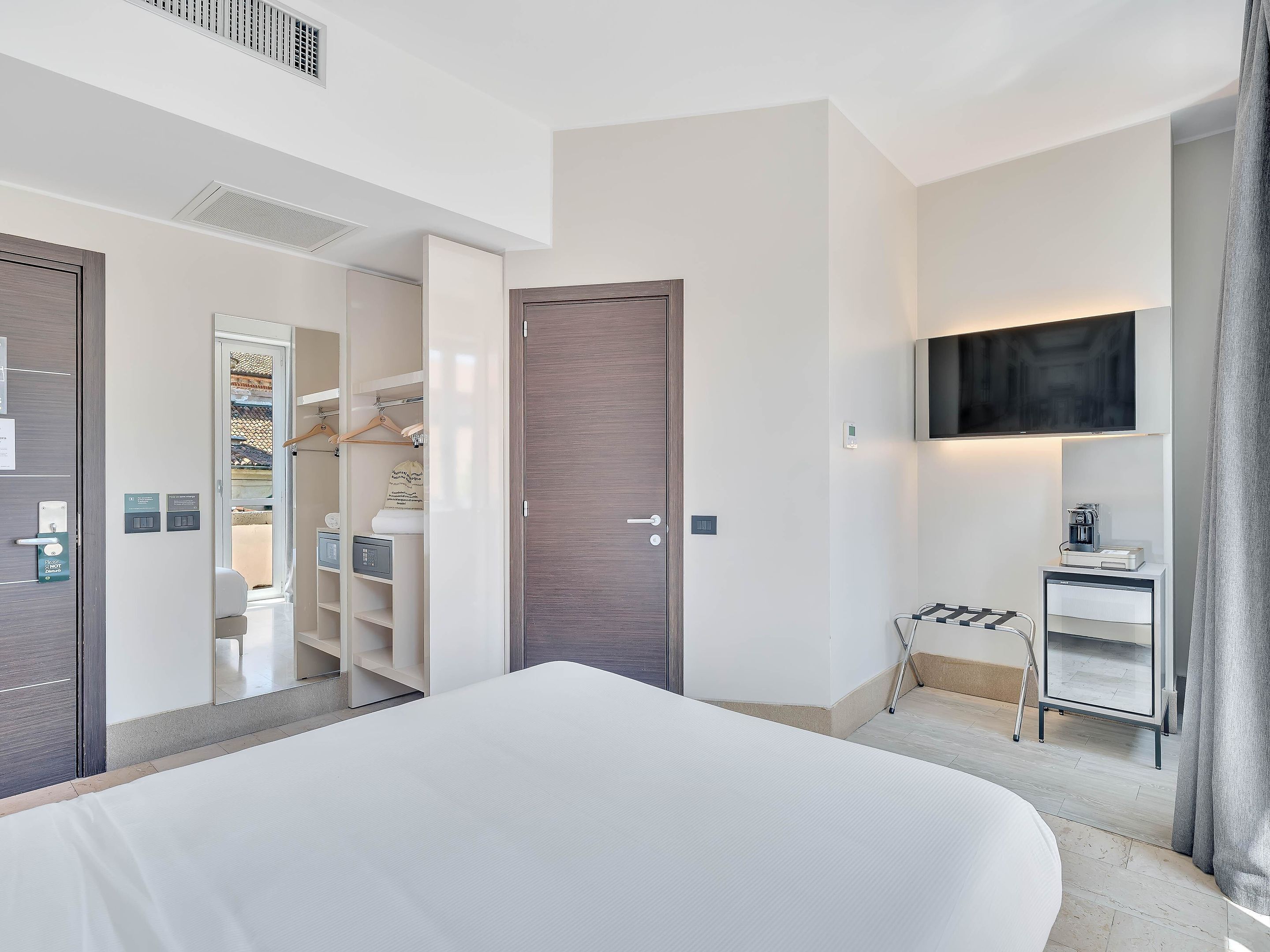 A modern hotel room with a large bed, a wardrobe, a television mounted on the wall, and a small bedside table with a coffee machine.